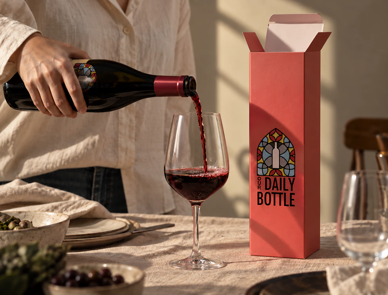 A person pouring red wine into a glass next to a tall coral-pink wine box branded 'OUR DAILY BOTTLE'