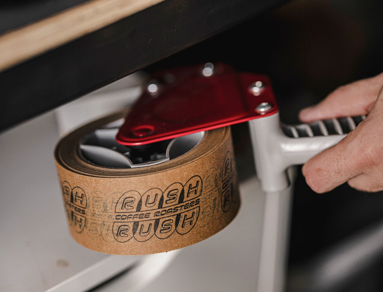 A hand using a tape dispenser with printed brown paper tape featuring the 'RUSH COFFEE ROASTERS' logo