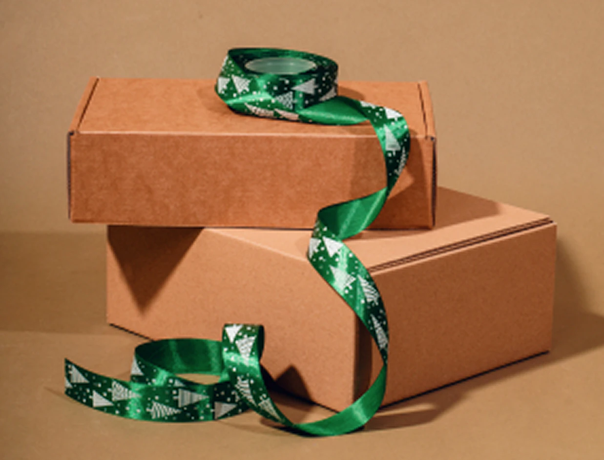 A roll of green satin ribbon printed with white Christmas tree patterns draped over stacked cardboard gift boxes