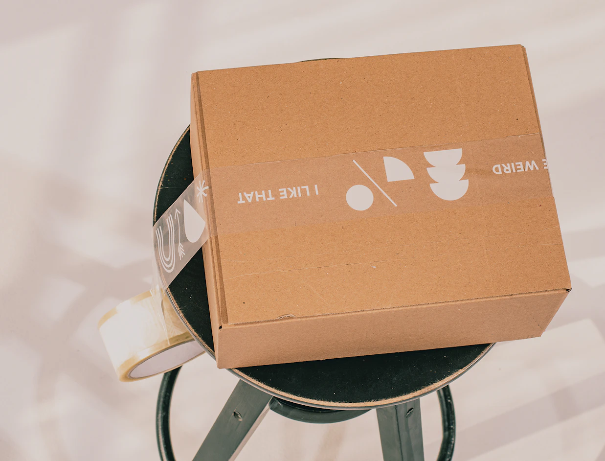 A closed cardboard box sealed with transparent tape printed with white graphics, sitting on a dark stool