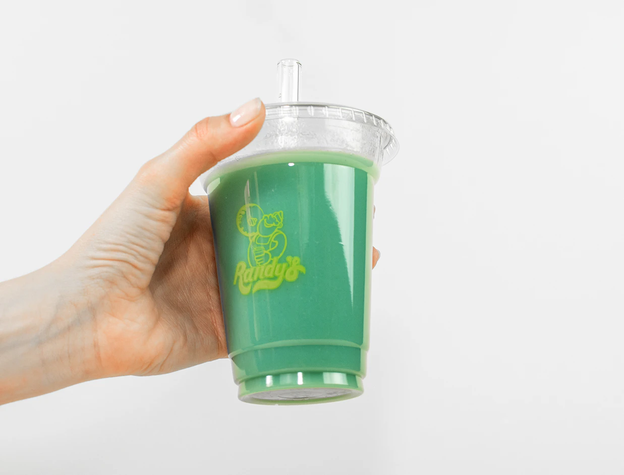 A hand holding a clear cup filled with green beverage and a glass straw, branded with the 'Randy's' logo against a light background