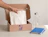 A hand taking a folded cream t-shirt out of an open 'PIXEL AGENCY' kraft cardboard box next to a water bottle and a blue notebook