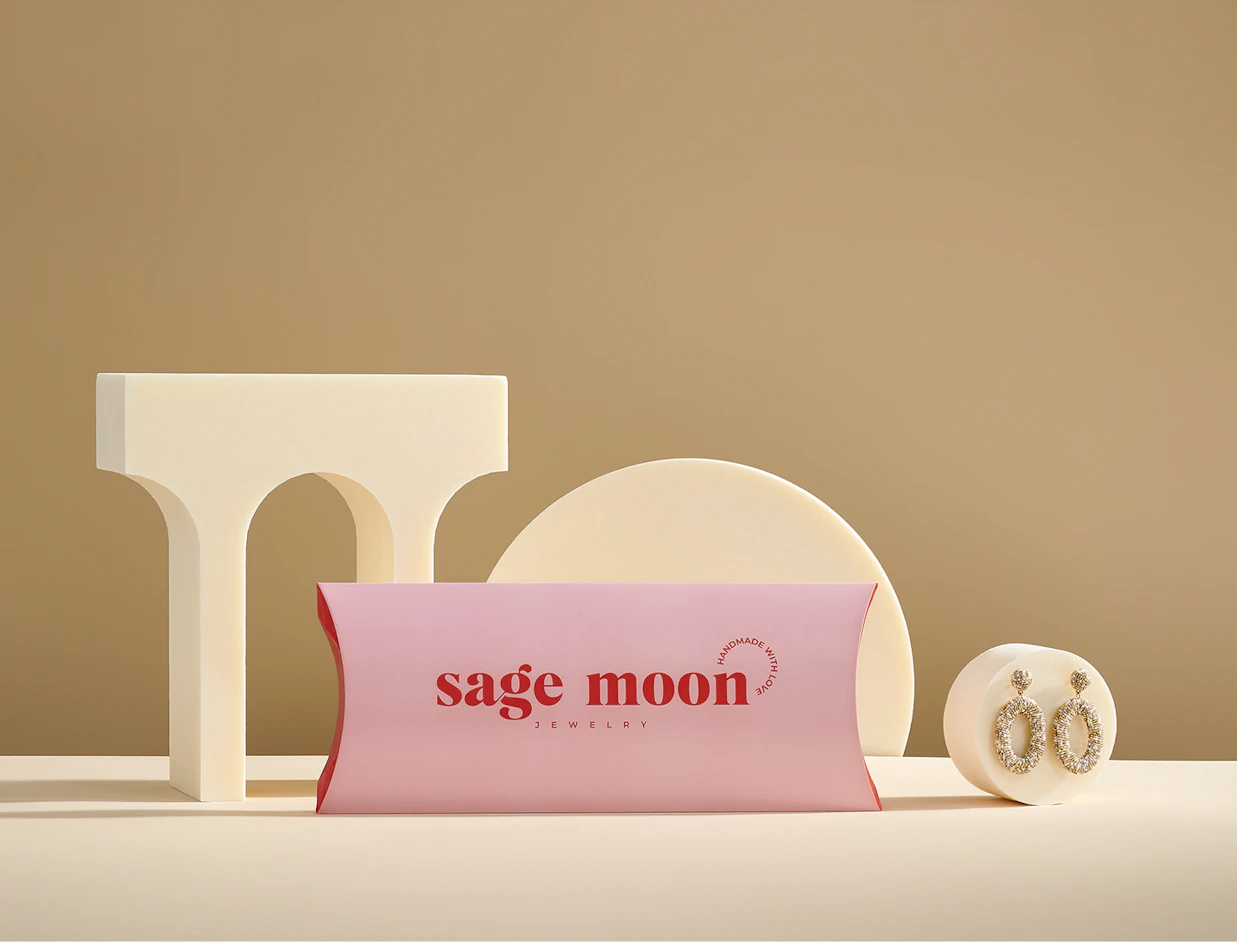 A pink pillow box branded 'sage moon JEWELRY' displayed on a beige background with geometric arch props and a pair of gold hoop earrings