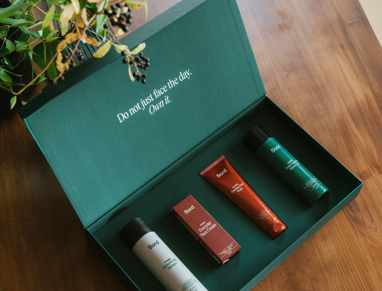An open forest green presentation gift box containing four Sord men's grooming products, with the inner lid printed 'Do not just face the day. Own it