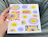 Hand holding a white sheet of stickers featuring smiling suns, clouds, flowers, and the word 'INNALE'
