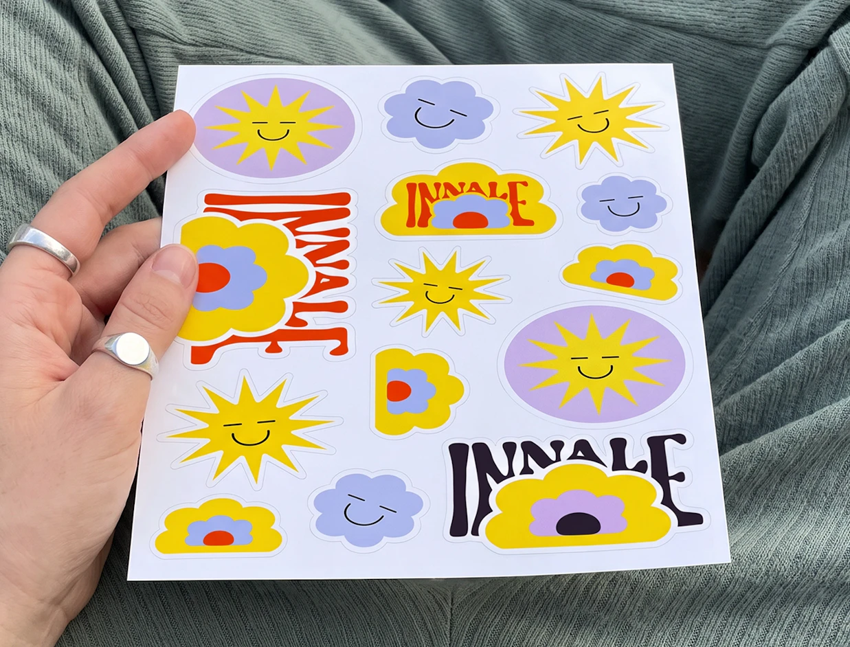 Hand holding a white sheet of stickers featuring smiling suns, clouds, flowers, and the word 'INNALE'