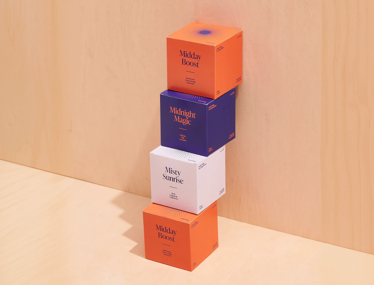 A stack of four colorful square candle boxes with product names 'Midday Boost', 'Midnight Magic', and 'Misty Sunrise' displayed against a light wood backdrop