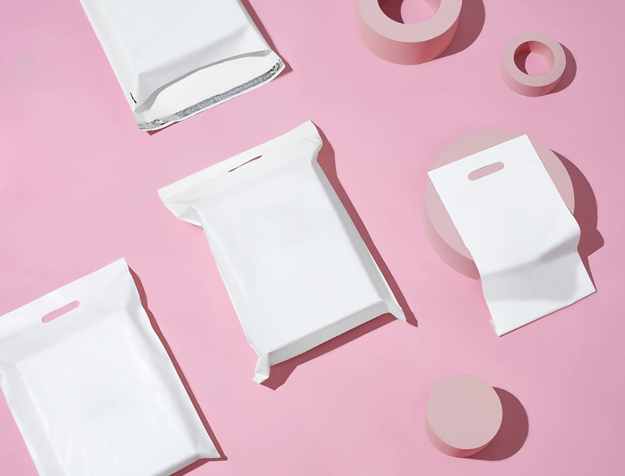 Flat lay of several blank white bubble mailers with die-cut handle cutouts arranged with pink geometric props on a pink background