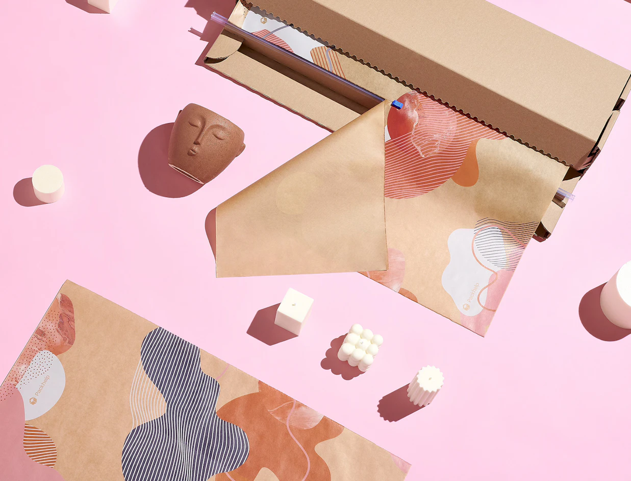 Crafting setup with rolls of abstract patterned wrapping paper, candles, and a face ceramic pot on a pink background