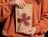 A person wearing an orange sweatshirt holding a kraft paper coffee pouch branded 'luma COFFEE' with a burgundy four-leaf clover print