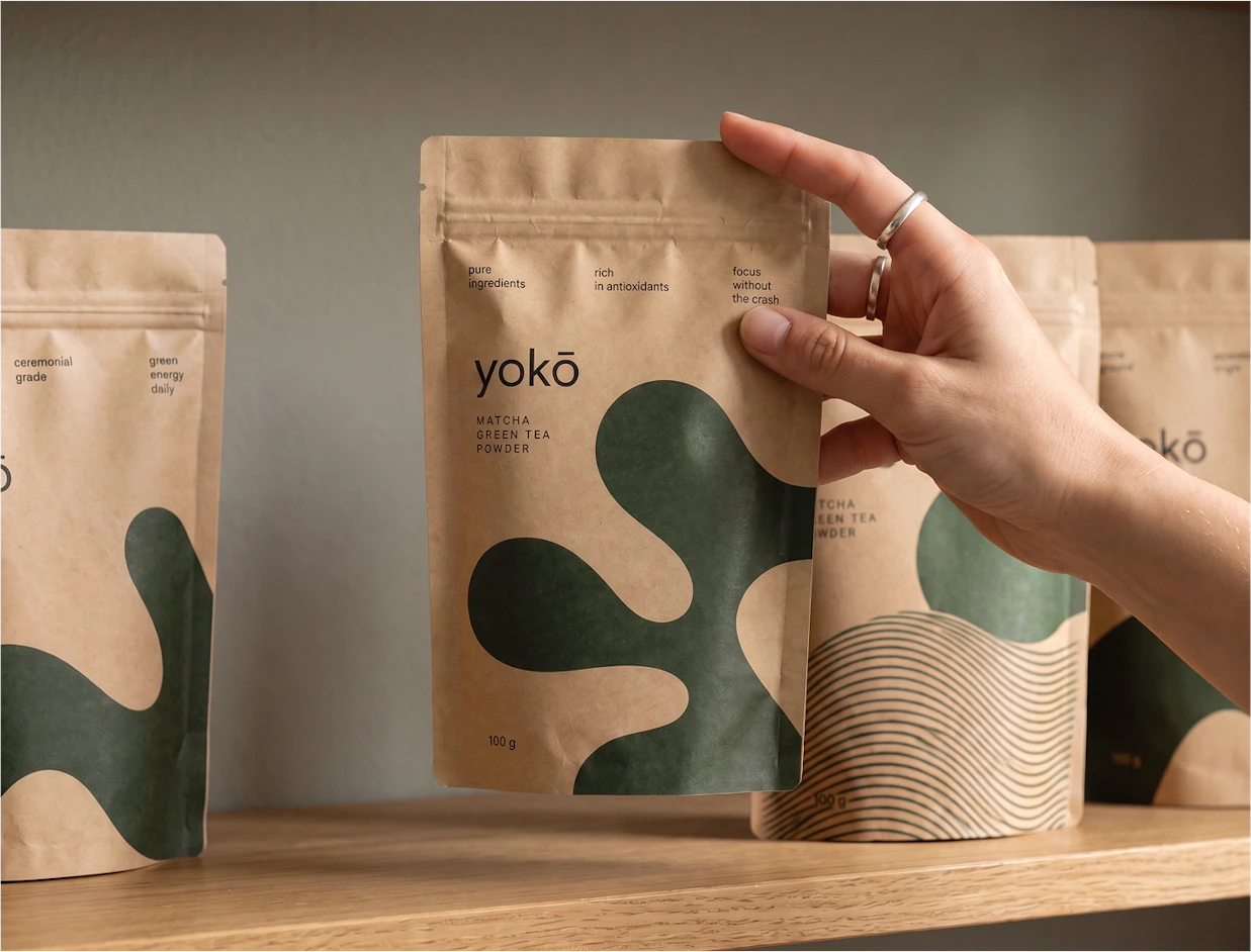 A hand reaching for a kraft paper stand-up pouch labeled 'yokō MATCHA GREEN TEA POWDER' with dark green abstract shapes, sitting on a wooden shelf