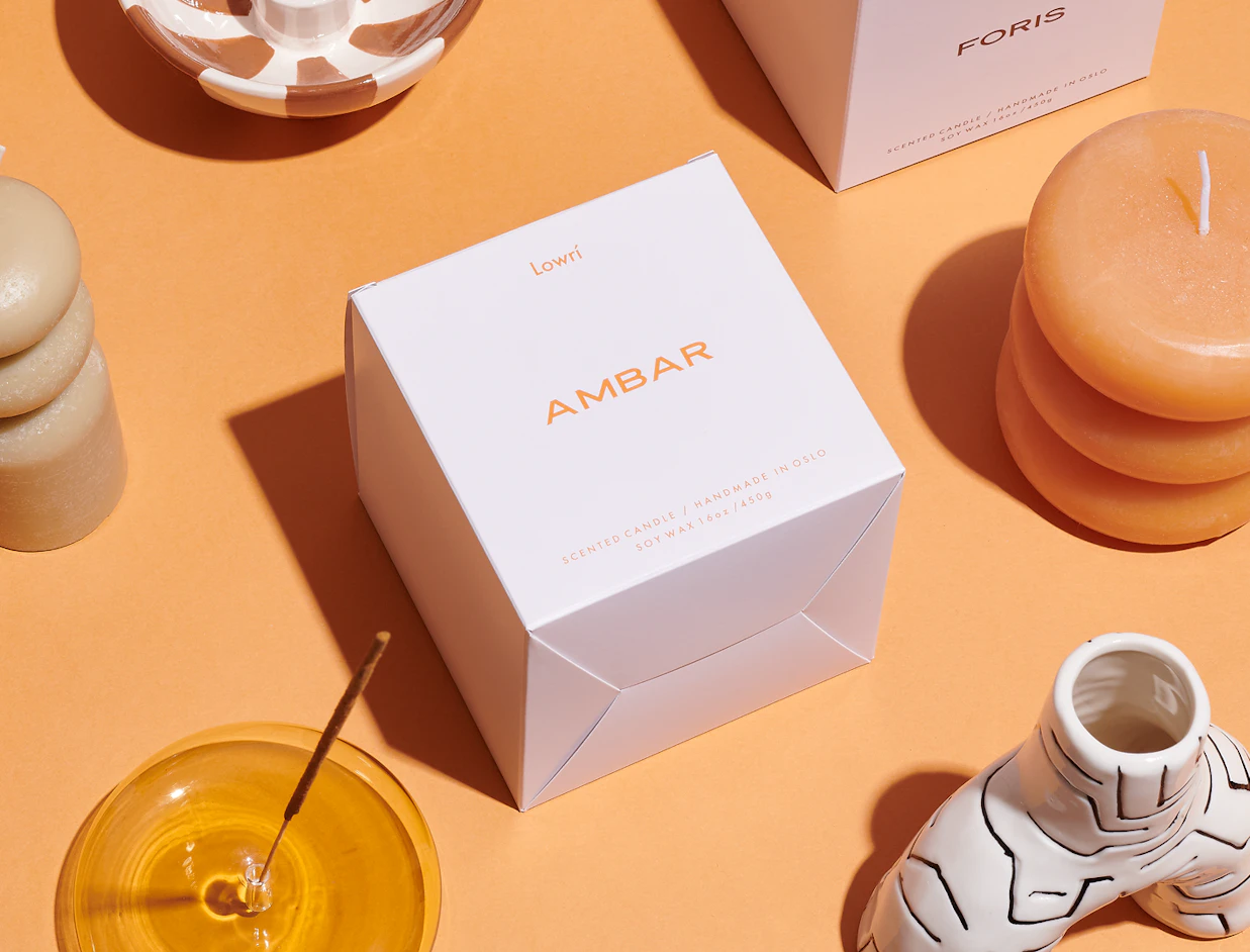 Top-down view of a white Lowrí 'AMBAR' candle box surrounded by stackable candles, an incense holder, and decorative ceramics