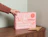A hand placing a pink mailer box with wavy red doodles and 'Hello, is it me you're looking for?' text on a wooden sideboard next to a brown envelope