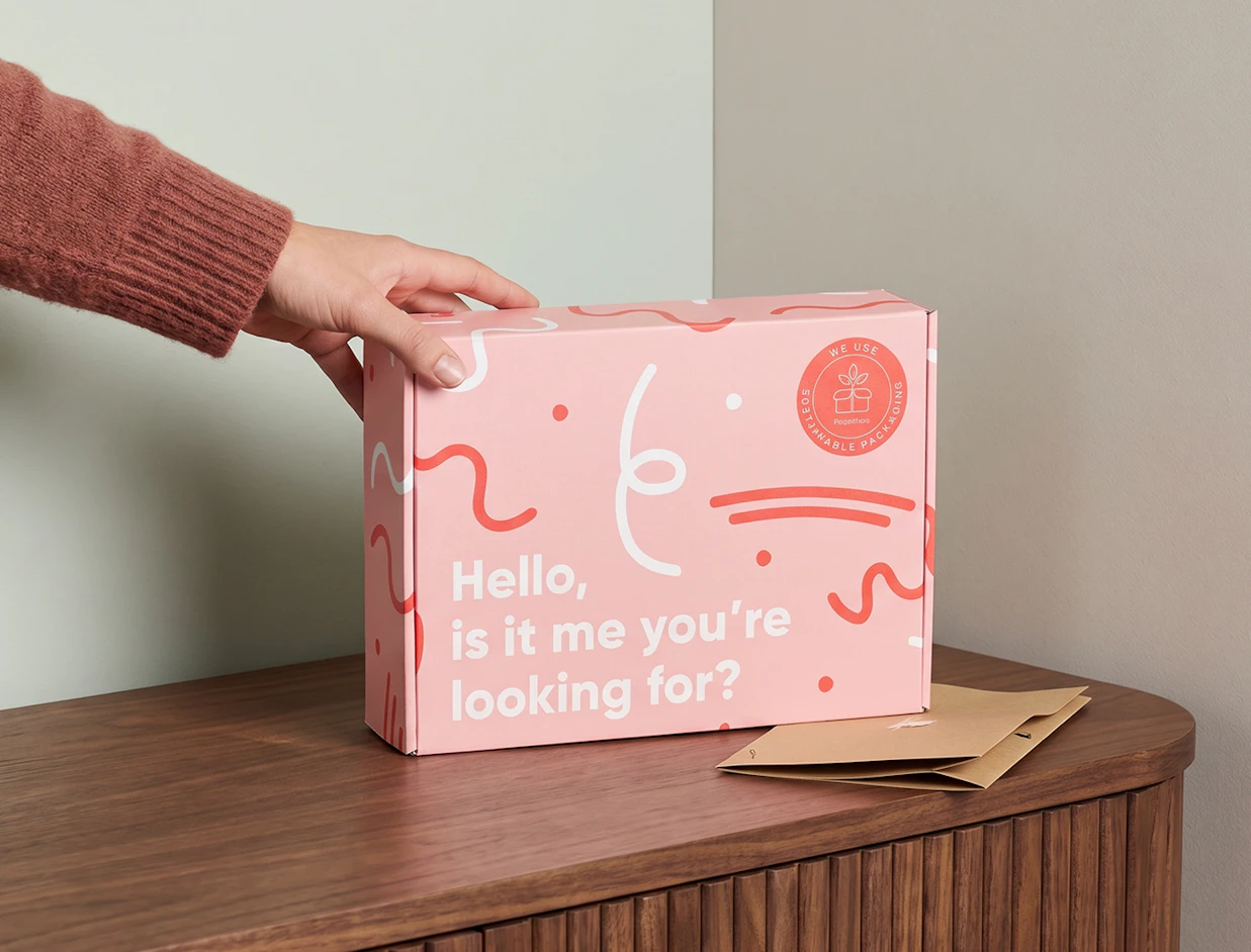 A hand placing a pink mailer box with wavy red doodles and 'Hello, is it me you're looking for?' text on a wooden sideboard next to a brown envelope
