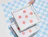 Hand holding a white pizza box featuring a black grid pattern and pink dots, resting on a stack over a blue checkered background