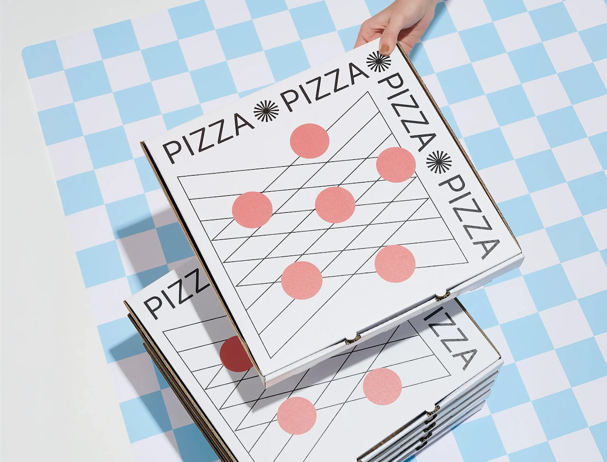 Hand holding a white pizza box featuring a black grid pattern and pink dots, resting on a stack over a blue checkered background