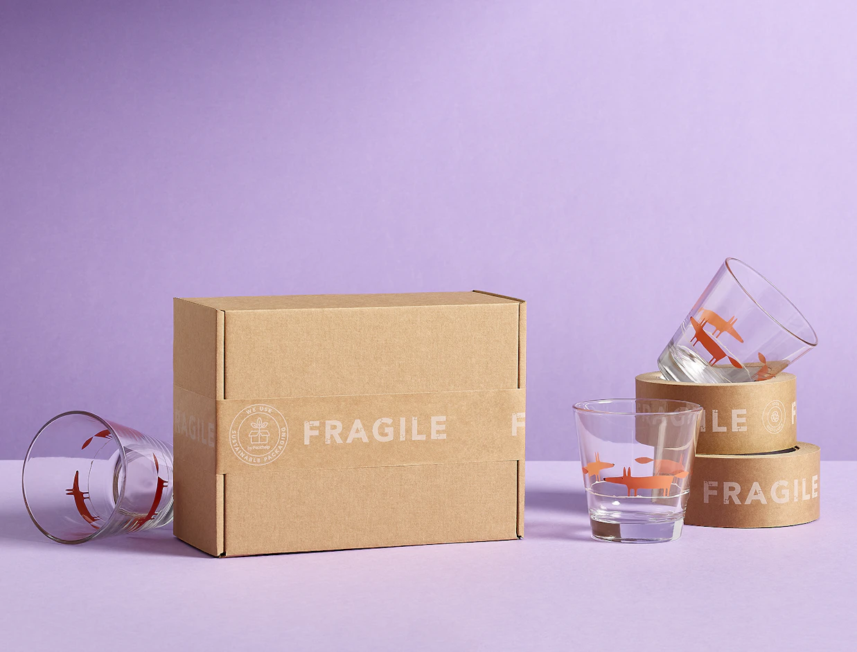 A brown cardboard box sealed with kraft paper tape printed 'FRAGILE', displayed on a purple background next to printed drinking glasses and rolls of tape
