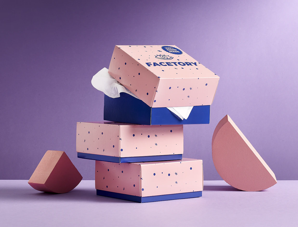 A stack of pink and blue dotted gift boxes branded 'FACETORY' surrounded by geometric foam shapes on a purple background