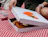 Hands opening a white pizza box printed with an orange tomato logo, revealing a fresh pizza on a red checkered picnic blanket