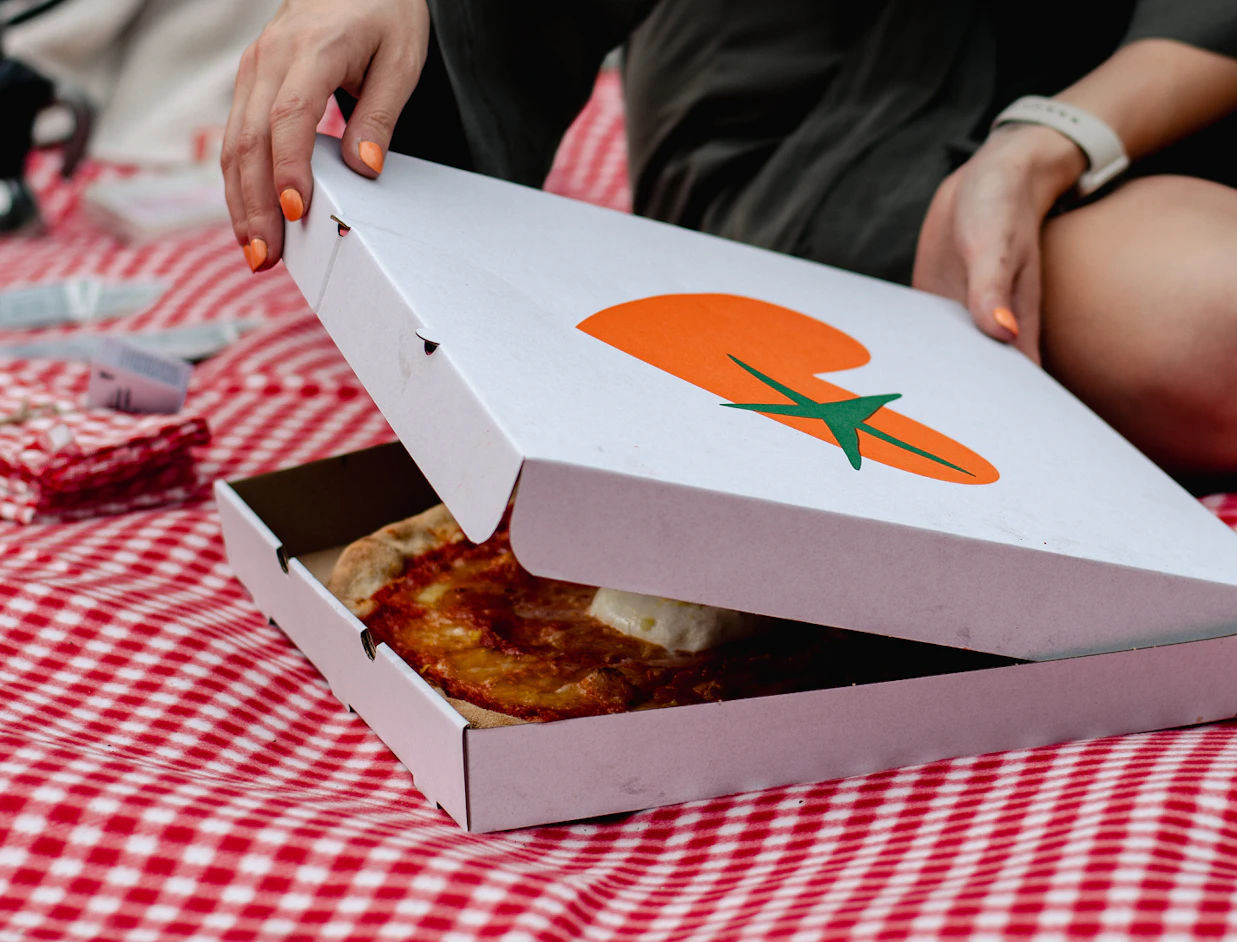 Hands opening a white pizza box printed with an orange tomato logo, revealing a fresh pizza on a red checkered picnic blanket