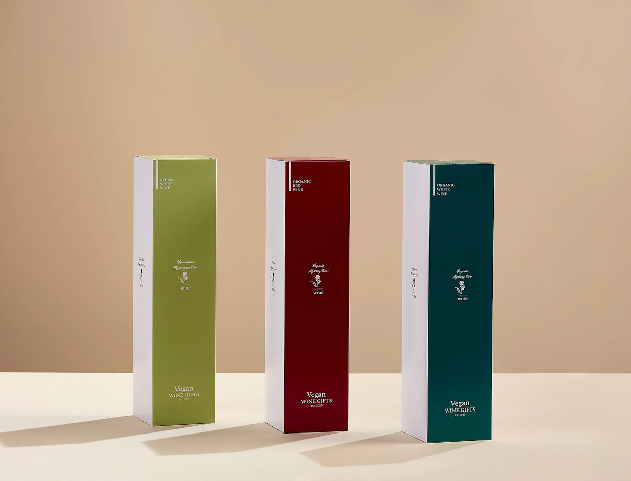 Three tall wine gift boxes in green, burgundy red, and dark green labeled 'Vegan WINE GIFTS' standing on a light surface
