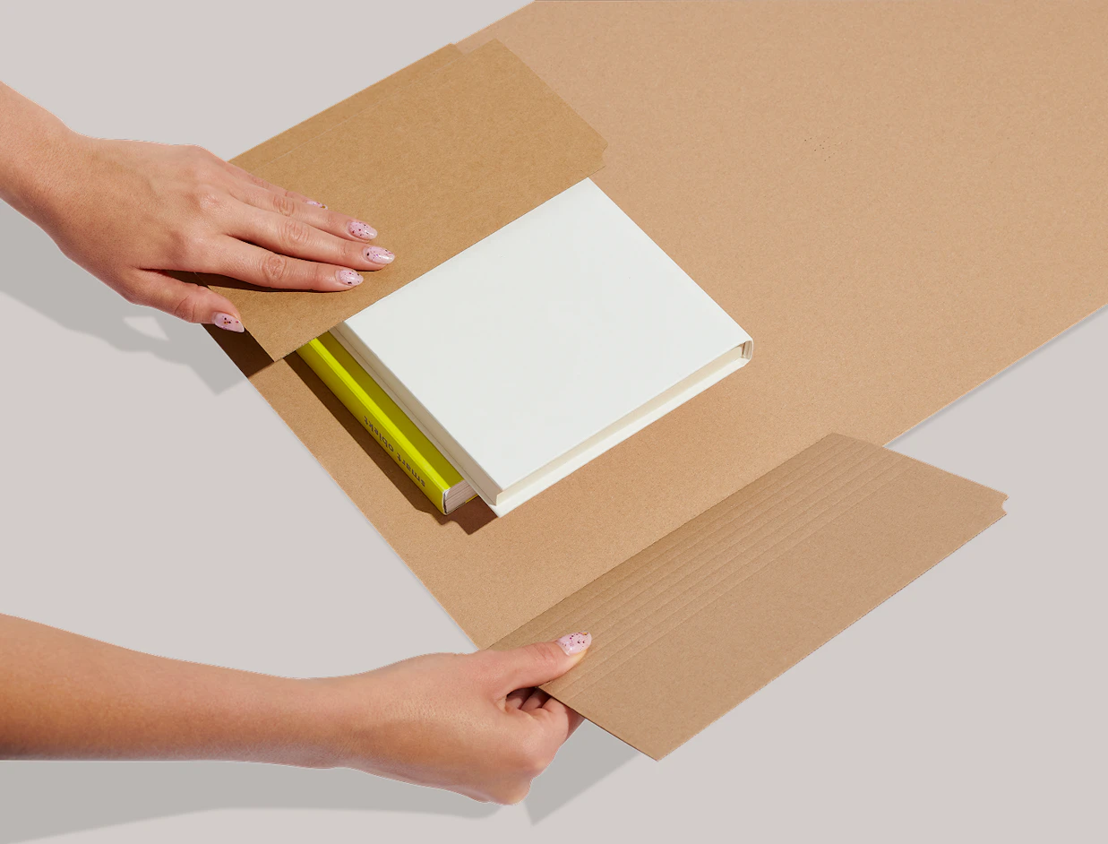 Hands wrapping books into an adjustable kraft cardboard book mailer packaging on a light background