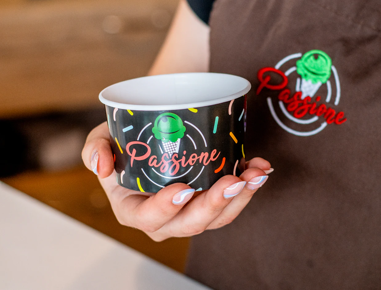 Close-up of a person wearing a brown apron holding an empty black paper ice cream cup with the 'Passione' logo