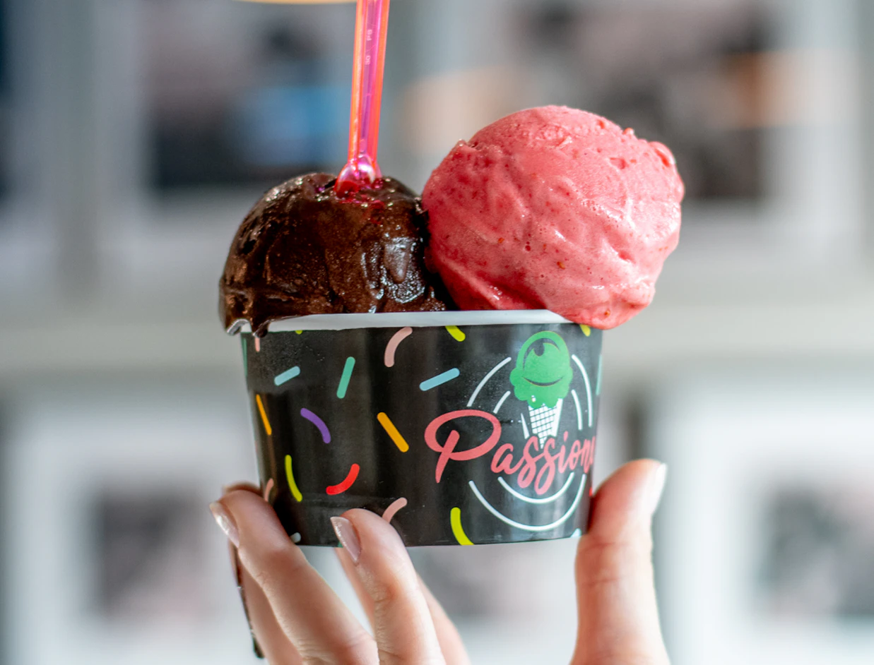 A hand holding a black paper cup labeled 'Passione' filled with scoops of chocolate and strawberry ice cream