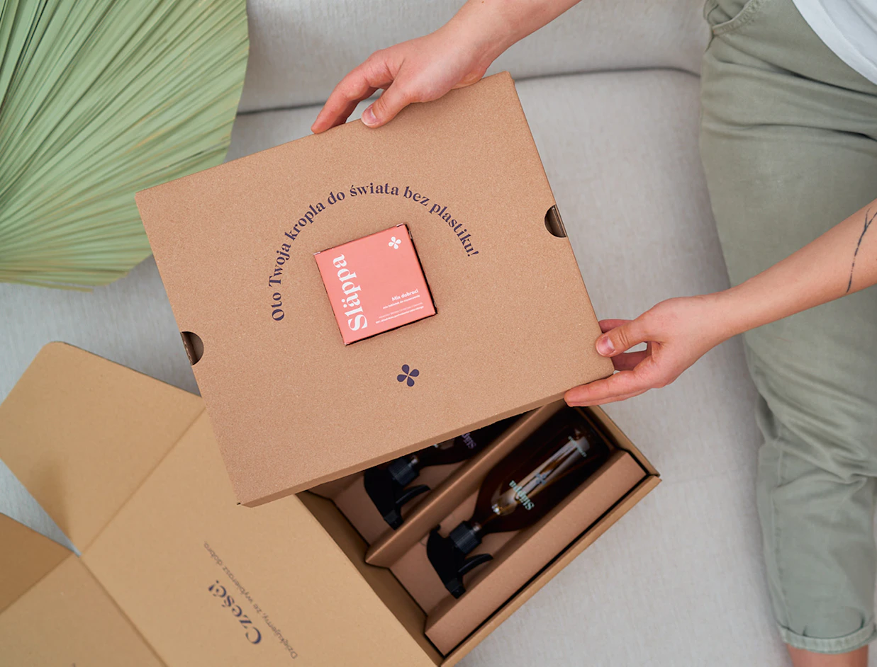 Hands unboxing a cardboard set with two glass spray bottles and a small pink Släppa box attached to the divider