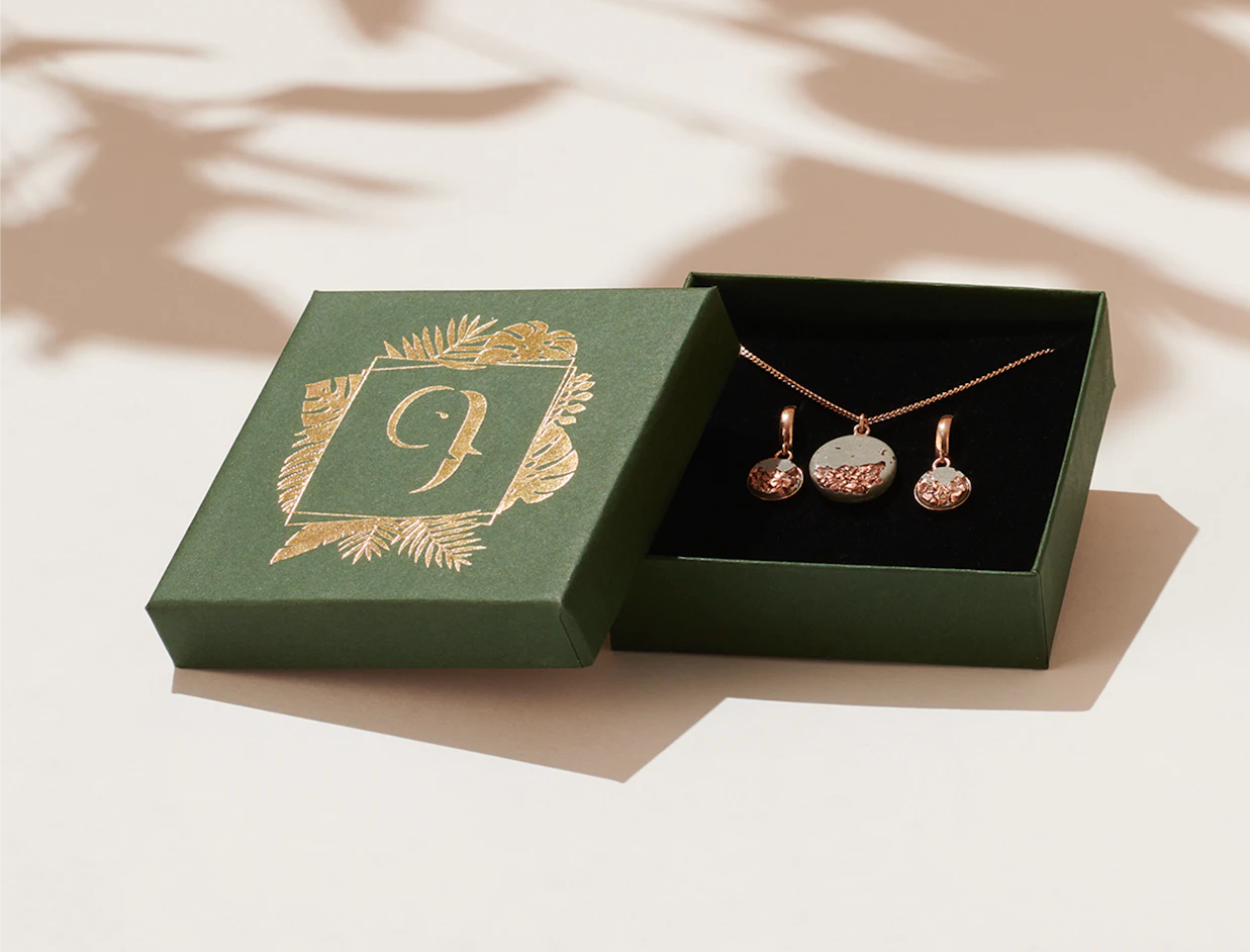 An open dark green jewelry box with gold foil palm leaf details displaying a necklace and matching earrings set