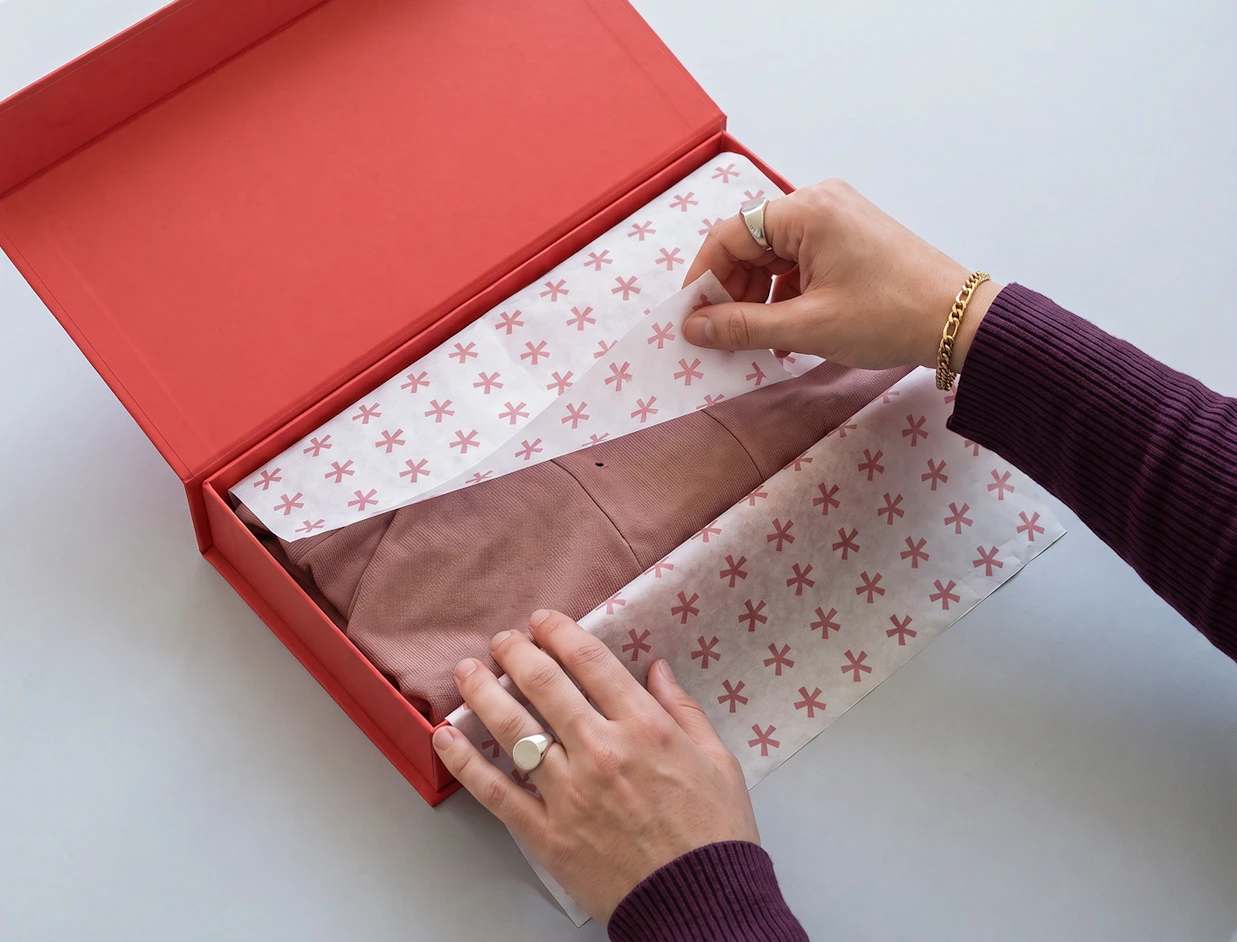 Hands carefully folding patterned tissue paper printed with pink star symbols over a garment in a red gift box