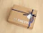 A brown cardboard gift box with silver metallic 'JJ EMLYN' typography, wrapped with a dark blue ribbon and a white gift tag on a light wood surface