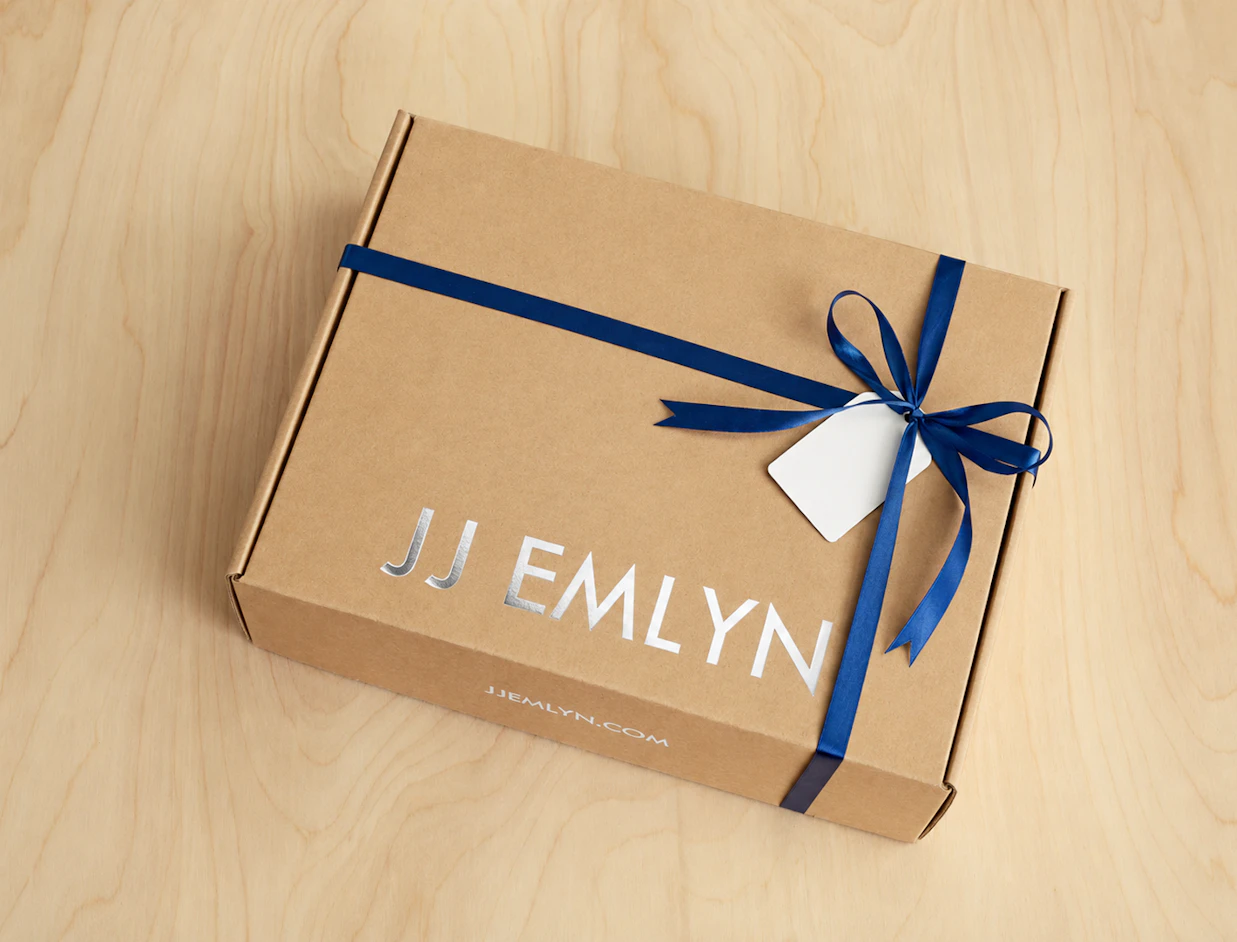 A brown cardboard gift box with silver metallic 'JJ EMLYN' typography, wrapped with a dark blue ribbon and a white gift tag on a light wood surface
