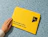 A hand resting next to a bright yellow cardboard envelope printed with 'cut the mustard' logo on a blue speckled surface