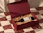 An open deep red presentation box holding a bottle of Pinot Noir on shredded paper over a checkered marble table
