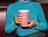 A person wearing a bright teal ribbed sweater holding a pink paper cup printed with red typography