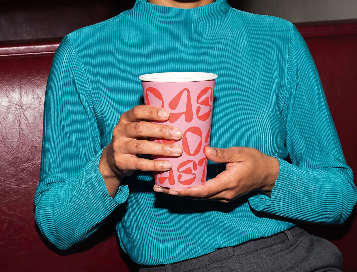 A person wearing a bright teal ribbed sweater holding a pink paper cup printed with red typography