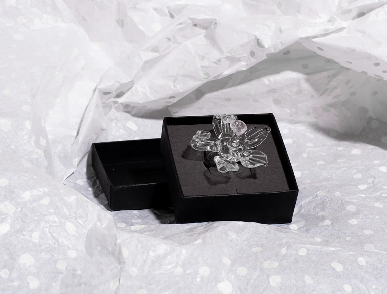 A small open black jewelry box holding a clear glass flower ring, placed on crumpled white polka-dot tissue paper