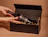 Hands with red nail polish carefully placing a wine bottle into a black gift box filled with paper filler
