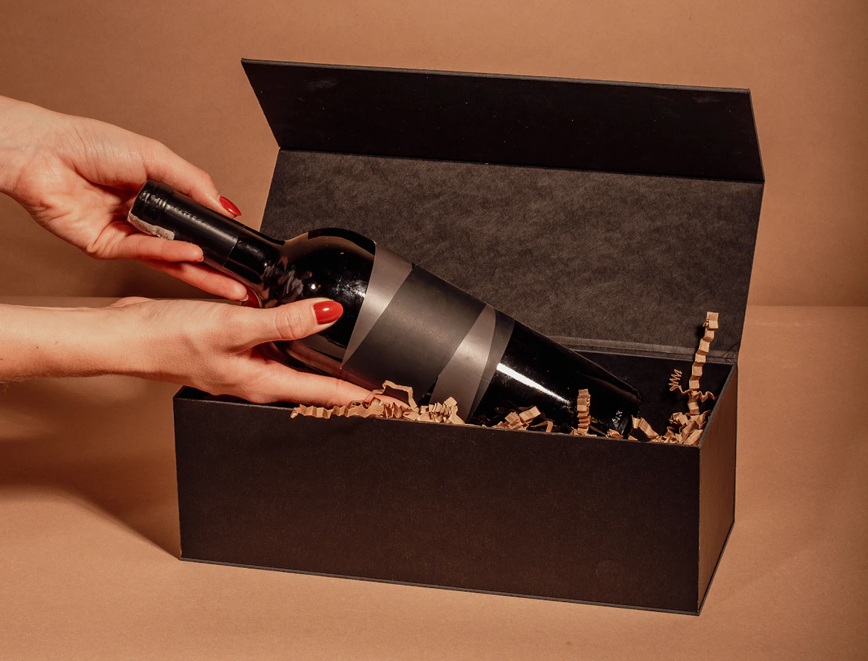 Hands with red nail polish carefully placing a wine bottle into a black gift box filled with paper filler