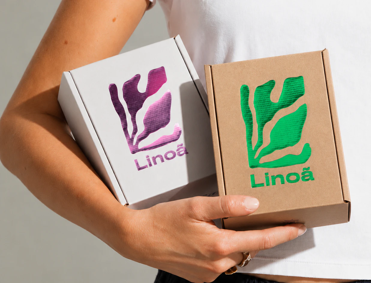 A person holding two small 'Linoã' shipping boxes, one white with purple metallic foil and one craft brown with green metallic foil