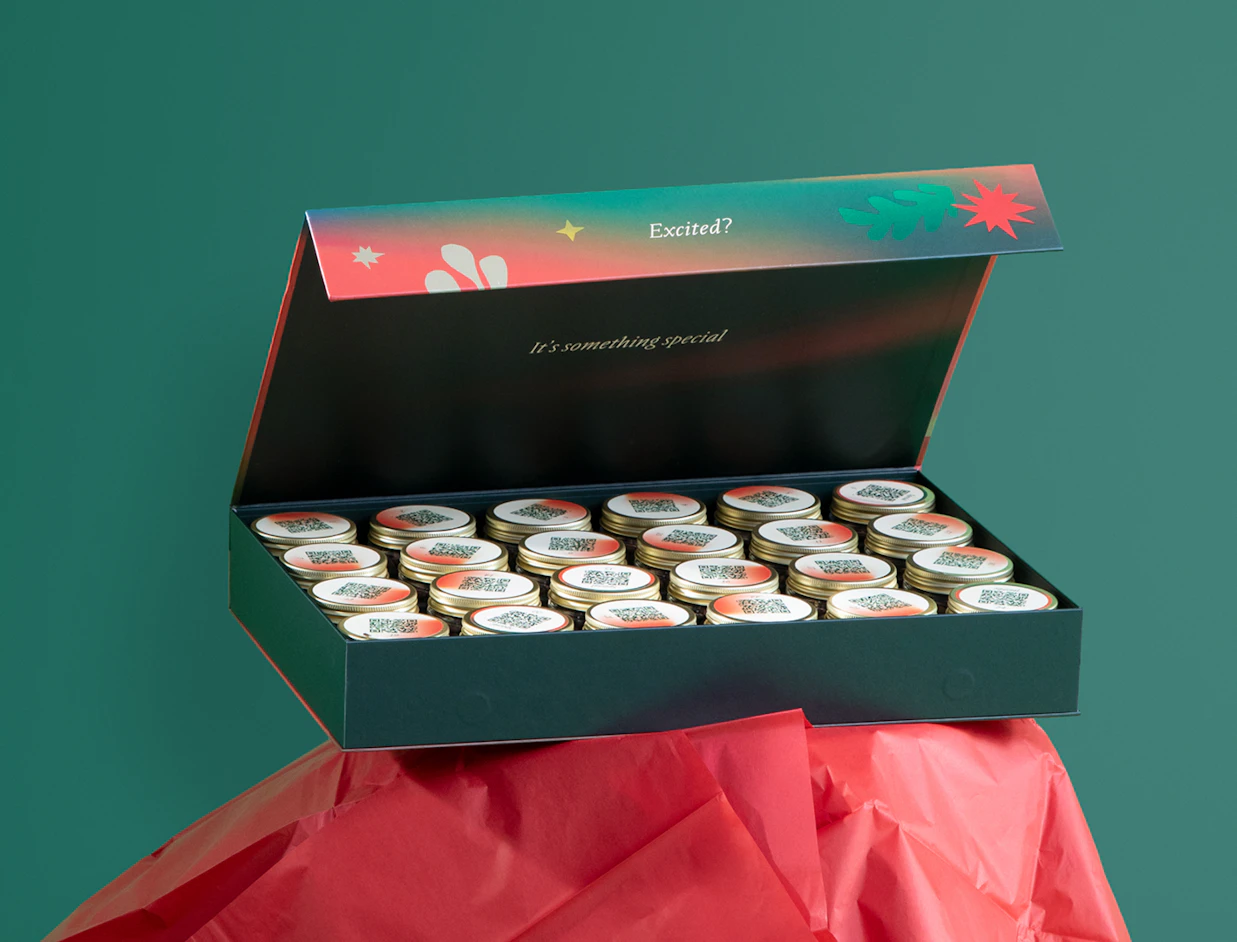 An open festive advent box filled with small glass jars with QR codes on red lids, set on red tissue paper