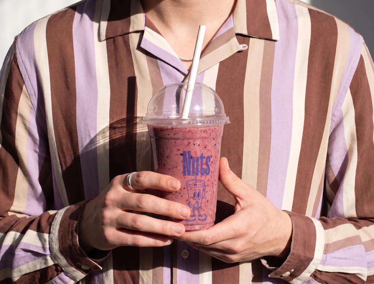 A person wearing a brown and purple striped shirt holding a clear plastic cup filled with a berry smoothie branded with 'Nuts'
