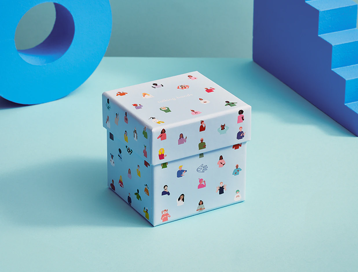 Light blue cube gift box decorated with colorful illustrated human figures, placed between blue geometric prop shapes