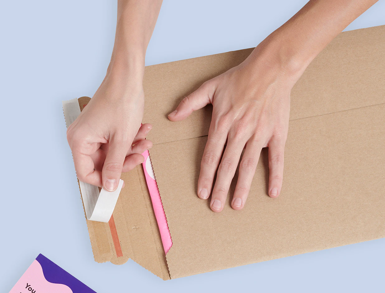 Top-down view of hands peeling the adhesive backing off a kraft cardboard envelope to seal it