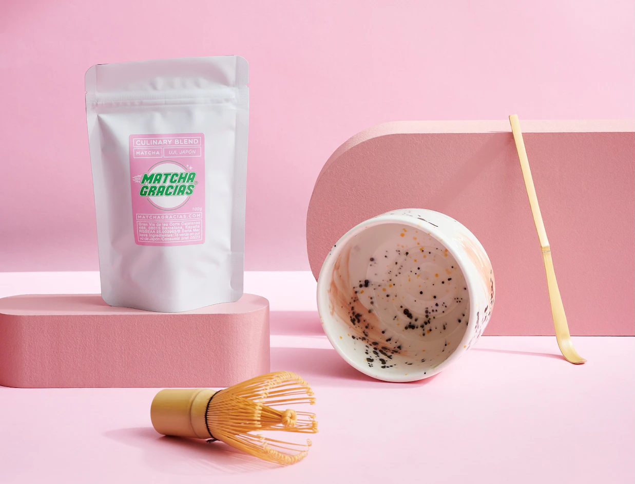 A white stand-up pouch labeled 'MATCHA GRACIAS' displayed on a pink background alongside a ceramic matcha bowl, bamboo whisk, and scoop