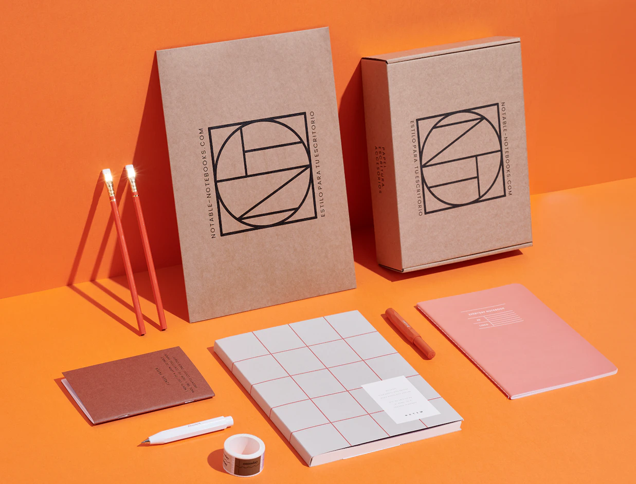 A collection of stationery items including notebooks, pens, and cardboard shipping boxes with 'NOTABLE-NOTEBOOKS.COM' branding on an orange background