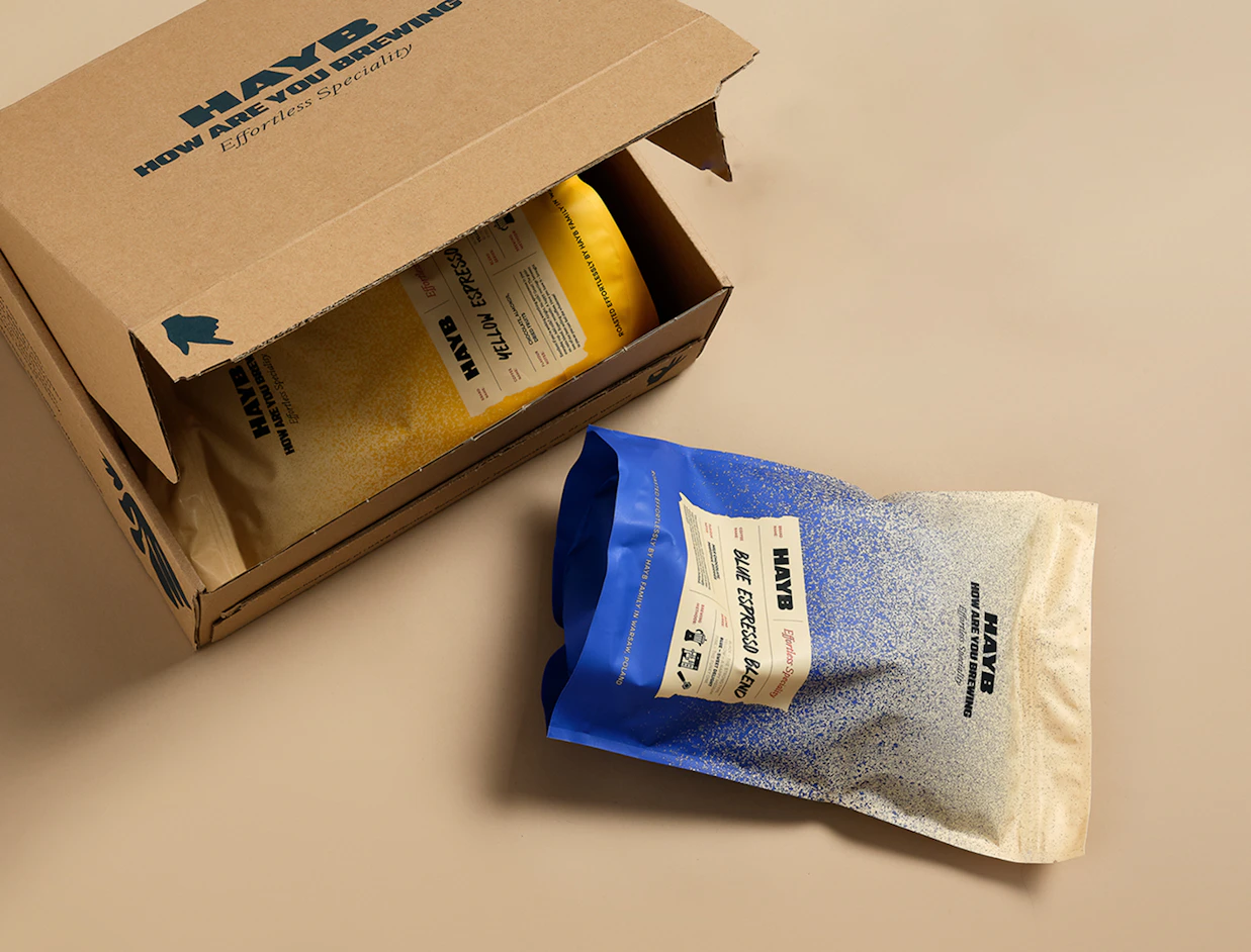 A blue coffee bag labeled 'HAYB BLUE ESPRESSO BLEND' lying next to an open cardboard shipping box containing a yellow coffee pouch