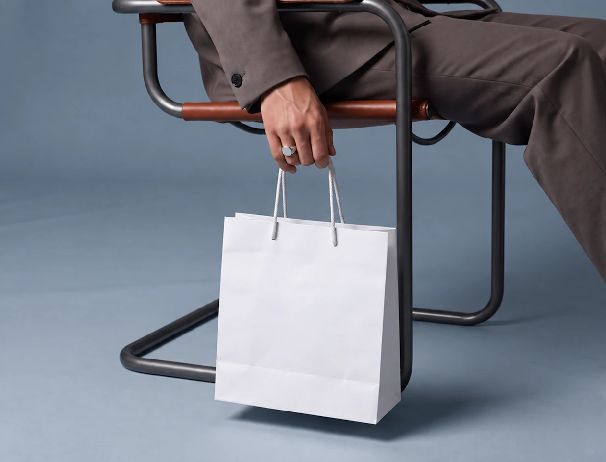 A person in a suit seated on a chair holding a blank white paper shopping bag with rope handles