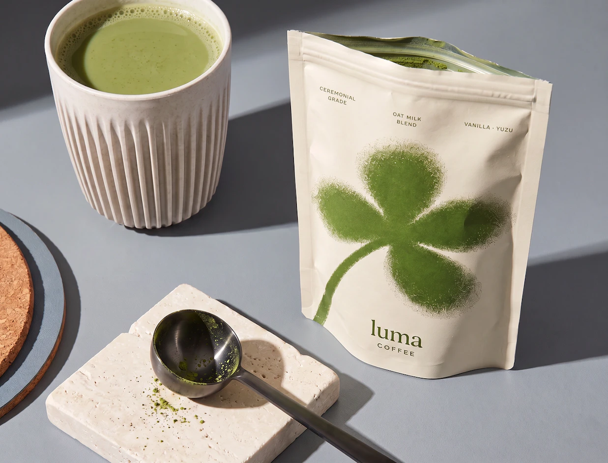 An open cream-colored stand-up pouch labeled 'luma COFFEE' next to a cup of matcha latte and a metal scoop